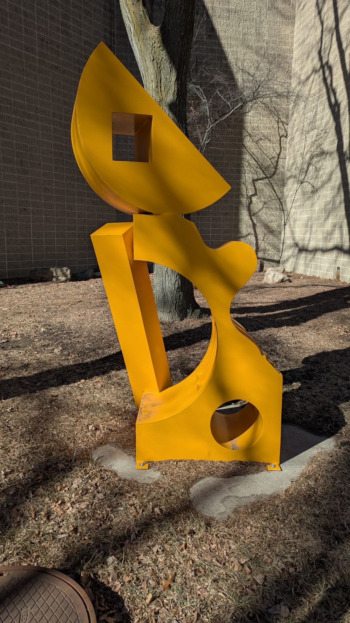 Bright yellow abstract metal sculpture with curved and geometric shapes displayed outdoors near a tree and building wall.