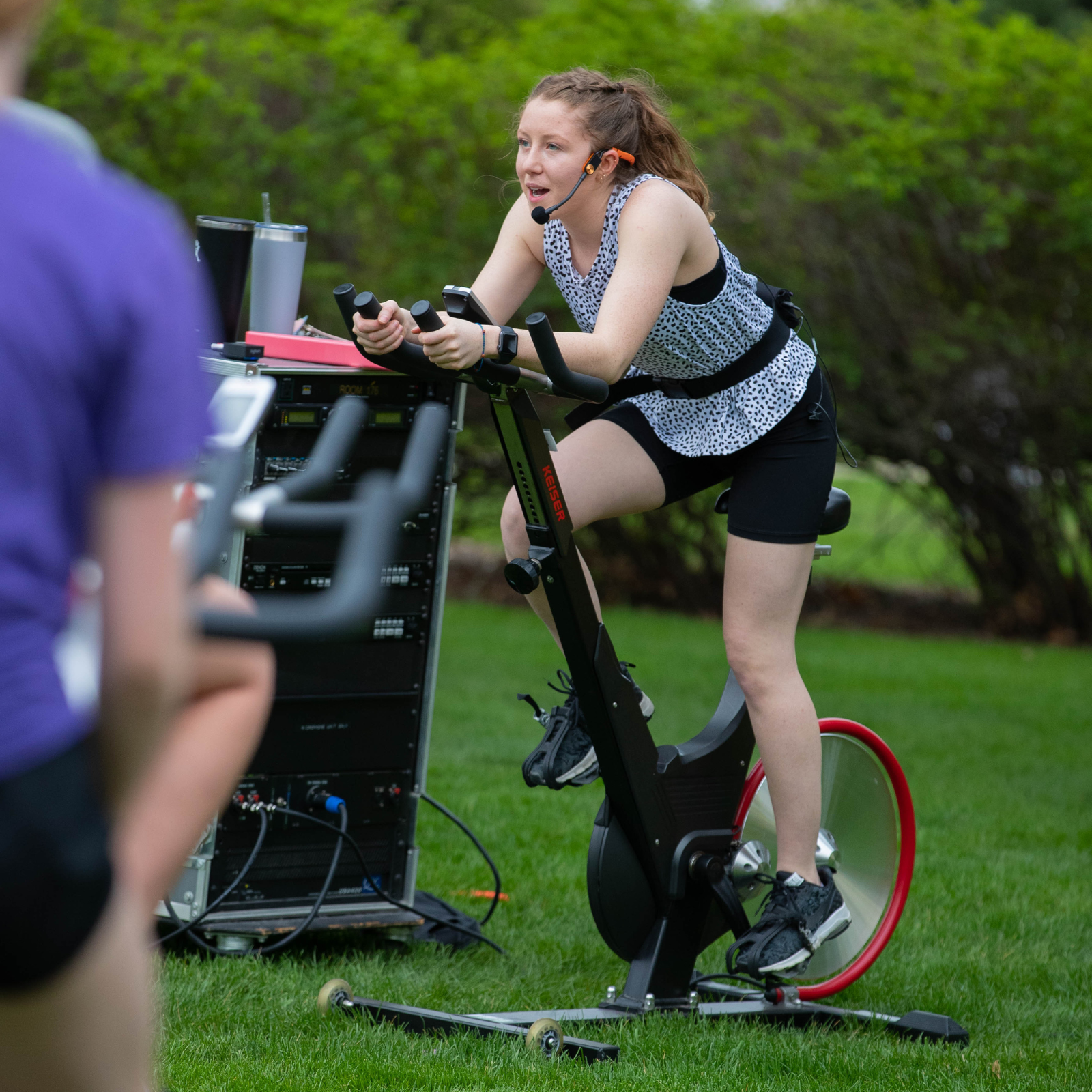 Person leading an outdoor cycling class on a stationary bike, wearing a headset and speaking while pedaling, with equipment and greenery in the background.